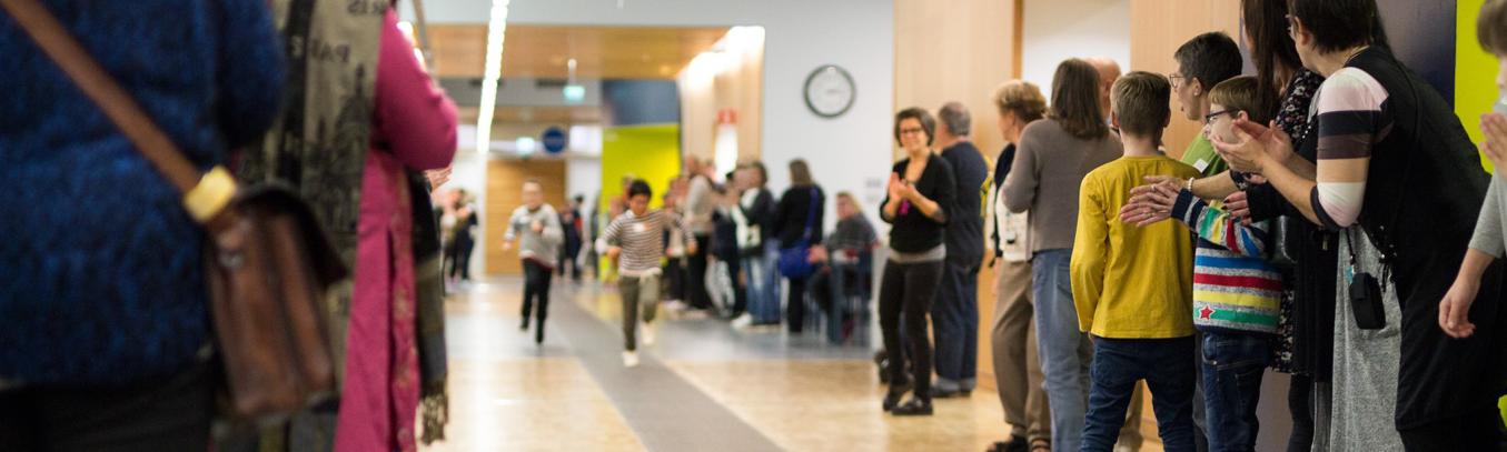 Pitkä, valoisa käytävä, jonka keskellä lapset juoksevat kilpaa. Käytävän molemmilla seinustoilla paljon tilannetta seuraavia ja taputtavia ihmisiä.